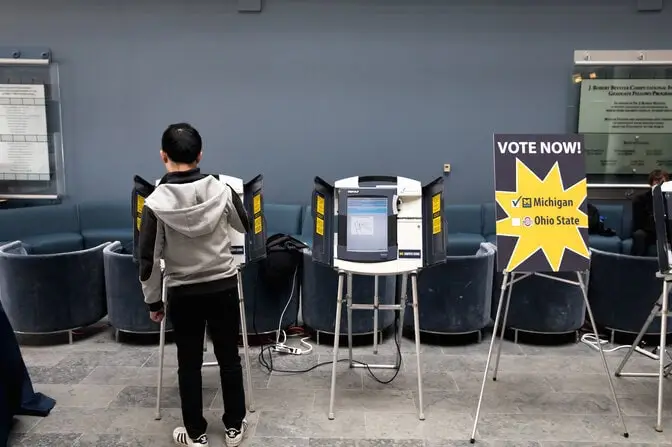  A sign next to two voting machines reads Vote now! with a checked box for Michigan and an empty box for Ohio State. The screen of one machine is visible but unreadable, while a voter blocks the view of the other.