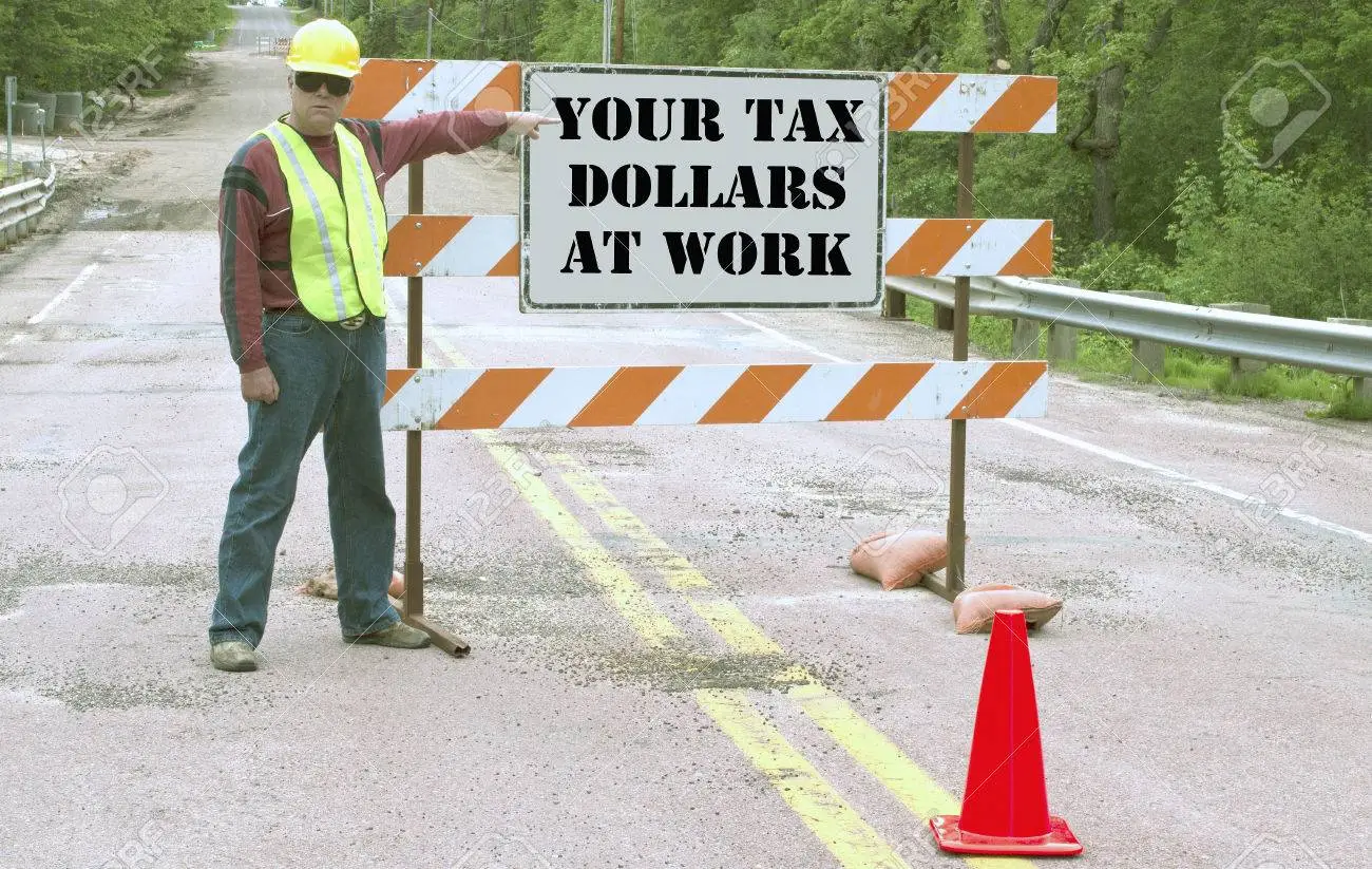24680107-construction-worker-points-to-a-road-sign-that-states-your-tax-dollars-at-work.webp